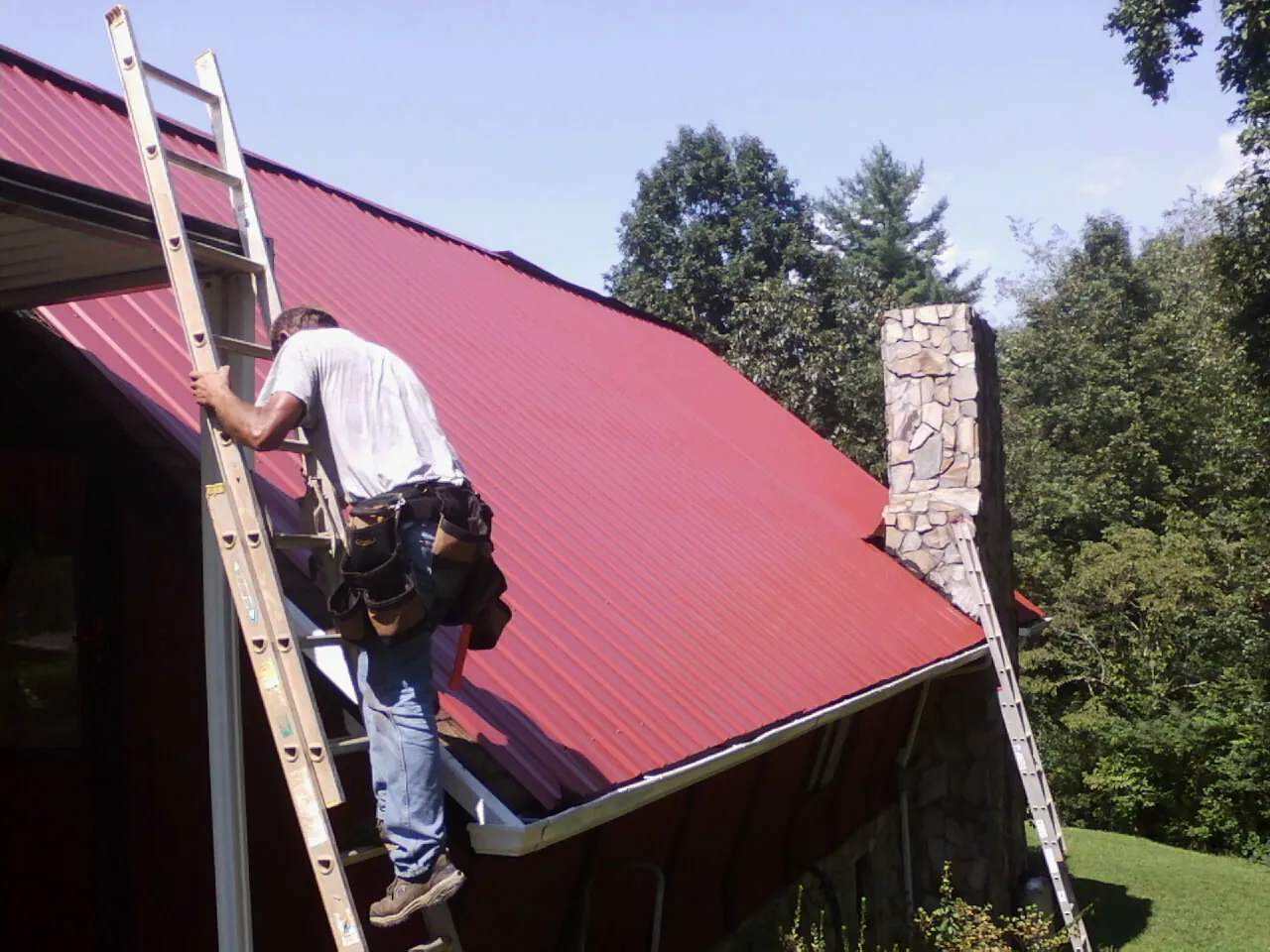 Roofer on a ladder beside a red metal roof and stone chimney — Soffit & Fascia Repair by Roofing Brothers in Panorama City